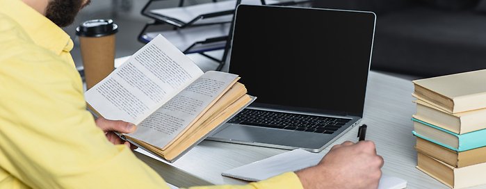 En person sitter och antecknar på ett papper med en laptop framför sig. I andra handen håller hen en bok. 